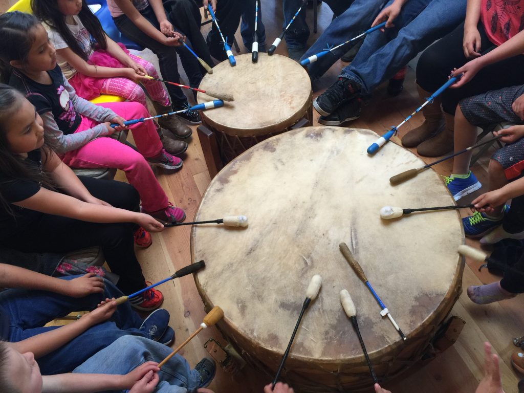 Native Blackfoot youth seated in a circle play a large drum.