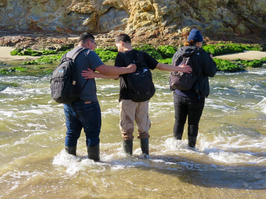 Three students help one another cross a river.