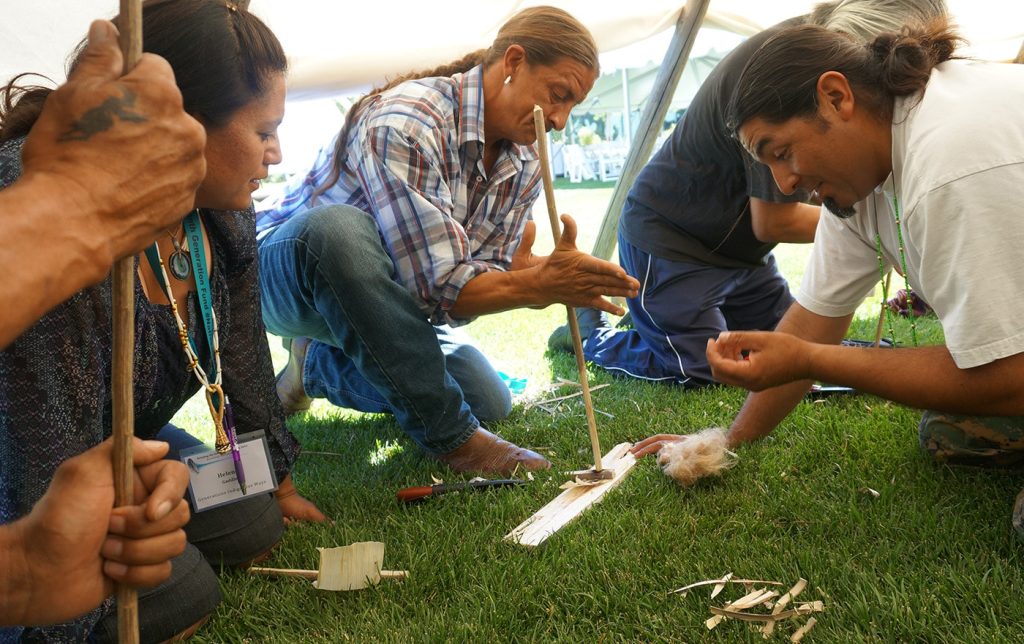 Indigenous people crouching in the grass practice fire-making with bow drills.