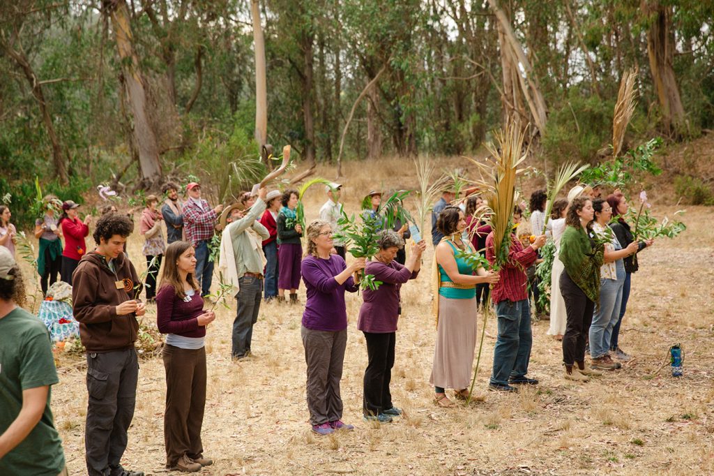 A group of people stand in a forest clearing, holding various offerings from nature.