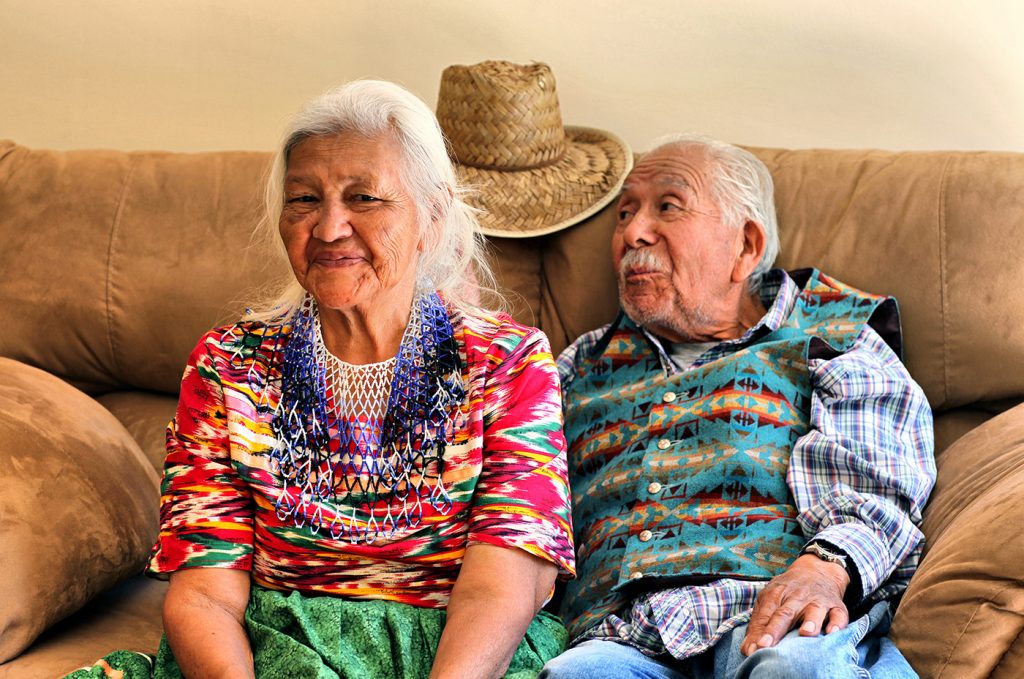 Two elderly people in traditional Zuni dress sit smiling on a couch.