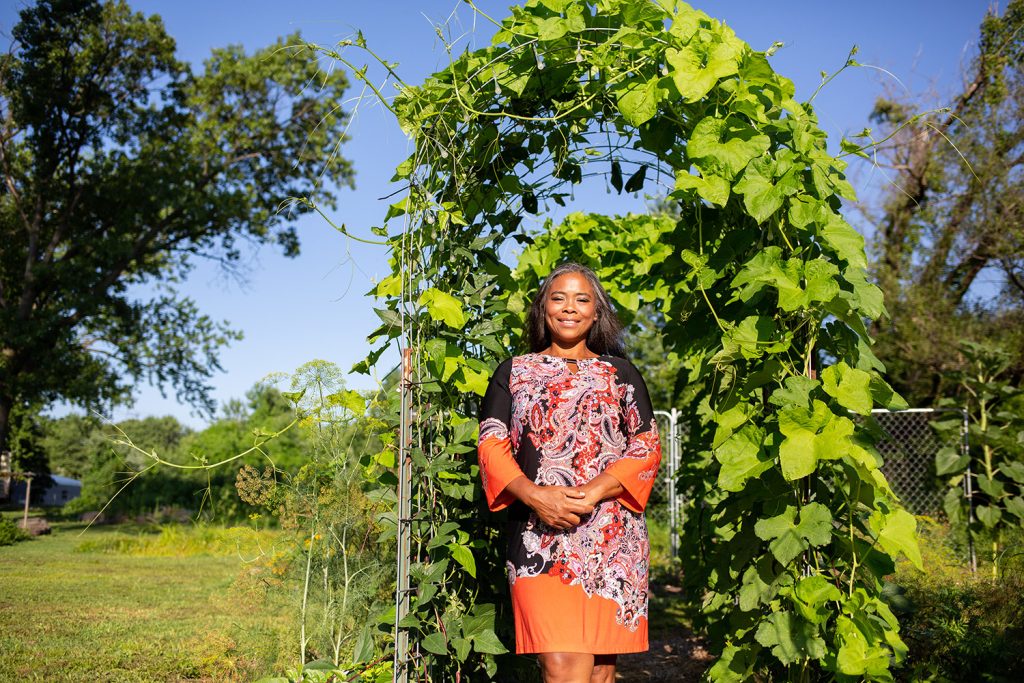 An African American woman standing in front of a leafy arbor in a colorful paisley dress at the Rustic Roots Sanctuary.