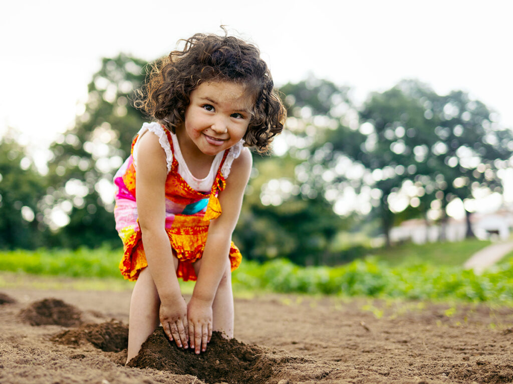 Little girl with curly hair and a colorful dress playing with the plowed land on her parents' farm.